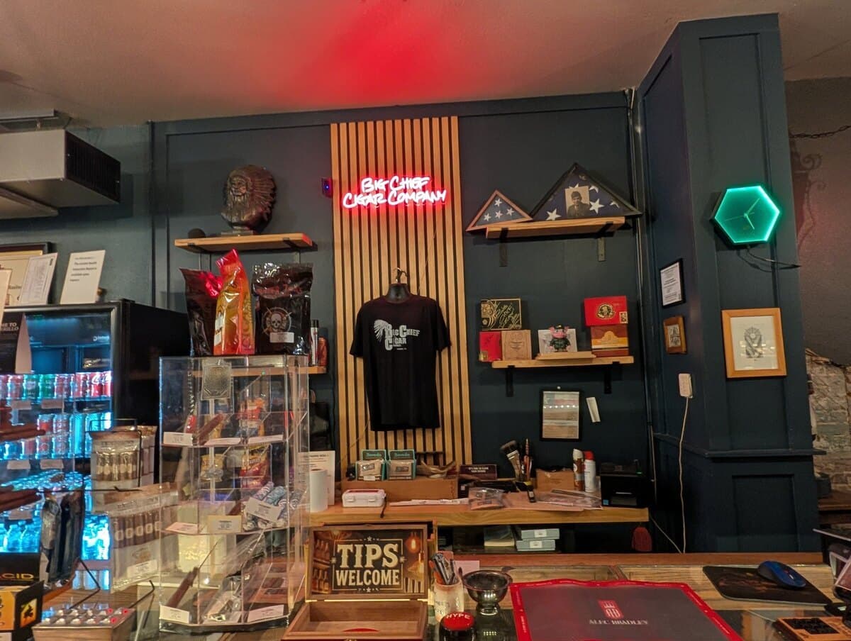 The front counter under a red neon Big Chief Cigar Company sign, with a shop T-shirt and folded flags on the wall